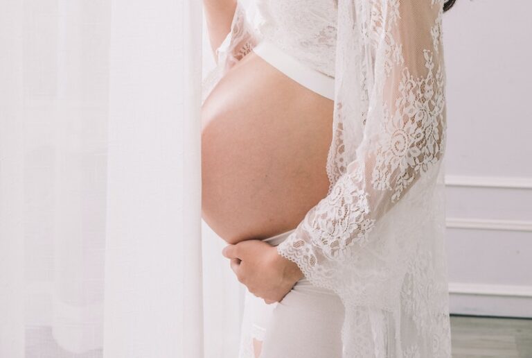 Happy young Asian pregnant female in white wear standing near curtain and looking at camera