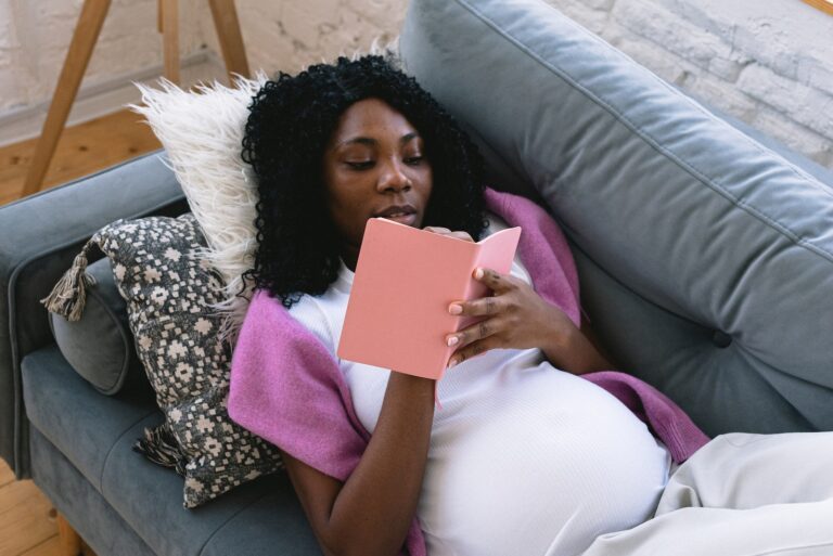 From above of focused pregnant African American female taking notes in notepad while resting on comfortable sofa in living room